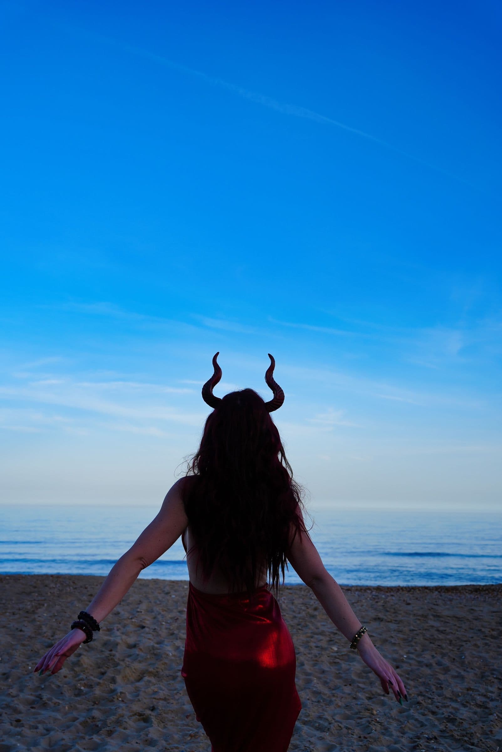 A figure with horns and long hair in a red dress walking across the sand under a deep blue sky