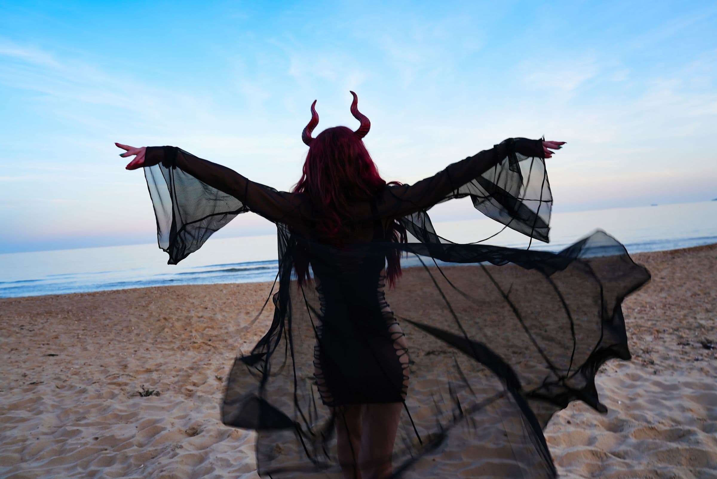 A horned figure seen from behind on the beach, sheer black sleeves spread like wings
