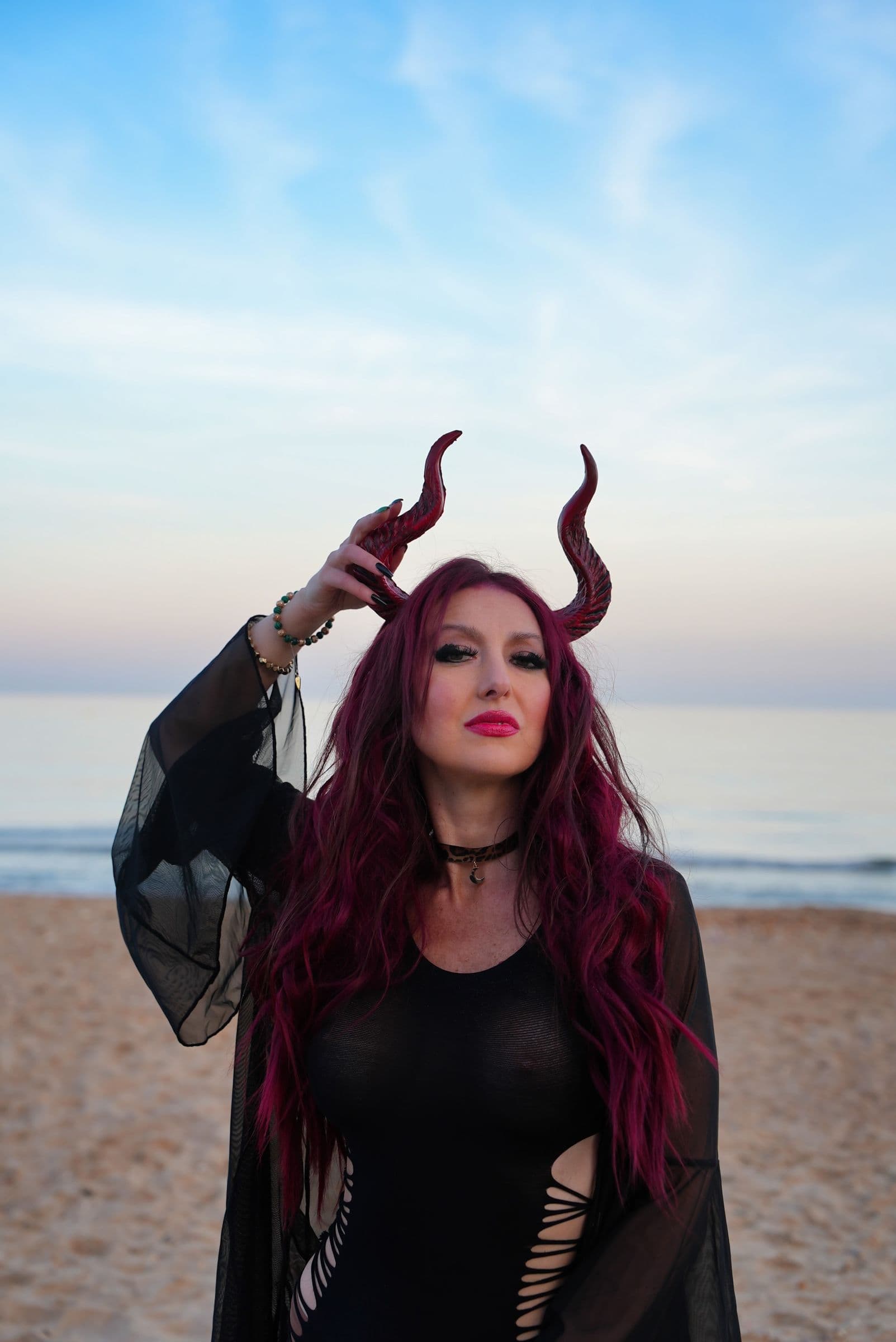 Yulia wearing horns and sheer black sleeves on the beach, one hand raised