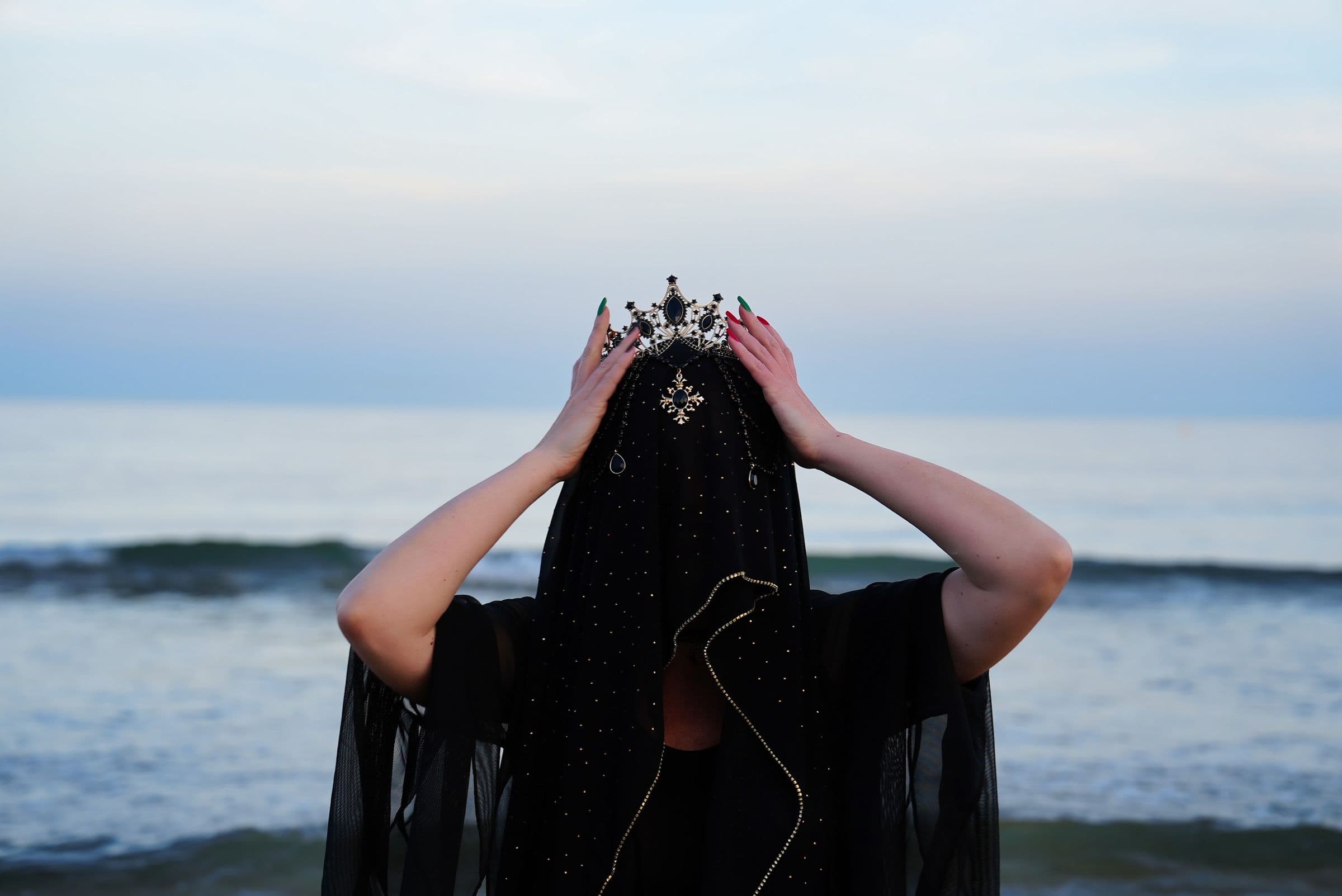 A figure in a jewelled crown and star-flecked black veil, hands raised, in front of the sea